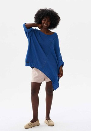 Blauwe oversized gebreide poncho met een diagonale zoom, geribbelde boorden en een textuurafwerking. Gecombineerd met beige shorts en instap geweven schoenen.