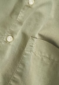 Camisa verde oliva hecha de un tejido ligero con una textura suave, con un diseño de botón y un bolsillo en el pecho.