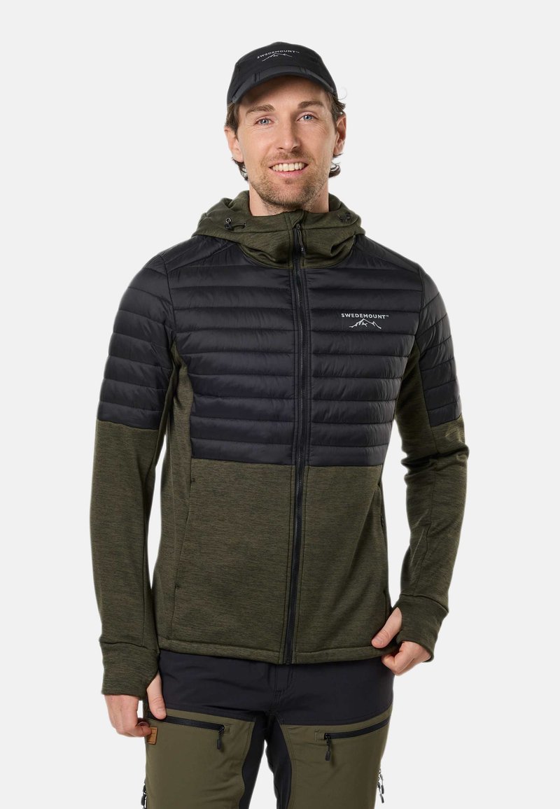 Homme souriant portant une casquette sombre, une veste à capuche bicolore verte et noire avec le logo Swedmount et un pantalon vert, debout face à l'avant.