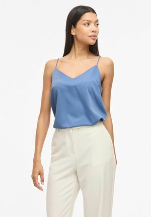 Femme portant un haut bleu sans manches et un pantalon blanc taille haute, regardant sur le côté avec un fond neutre.