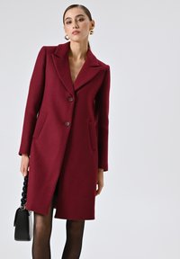 Bordeaux wollen mix jas met een geknipt kraag, twee voorzakken en knoopsluiting. Model houdt een zwarte handtas met een dikke strap vast.