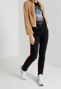 Krótka kurtka z tan fałszywego futra, czarne dżinsy z czerwonymi bocznymi paskami, czarna koszulka z grafiką, białe tenisówki z niebieskimi akcentami.