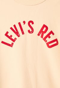 Sweat-shirt couleur crème présentant des lettres rouges audacieuses et usées formant l'inscription "LEVI'S RED" en arc de cercle. Tissu lisse et doux.
