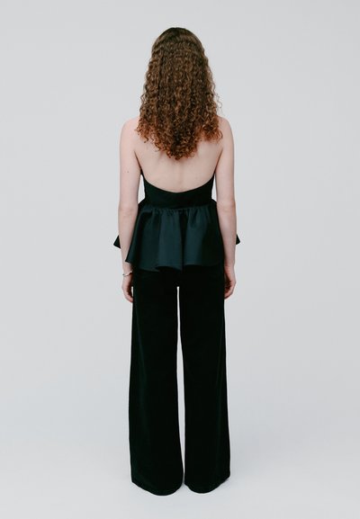 Femme aux cheveux bouclés portant un top peplum noir sans bretelles et un pantalon à jambes larges, debout de dos devant un fond uni.