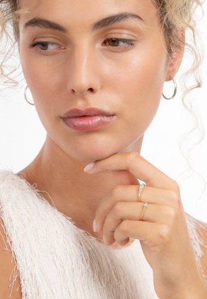 Close-up of a person with textured white top, wearing small gold hoop earrings and a delicate gold ring with a diamond accent.