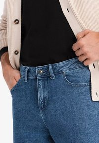 Persona vistiendo jeans azules, camiseta negra y cárdigan beige, ajustando la cintura de los jeans con una mano en el bolsillo y la otra tirando de la cintura.