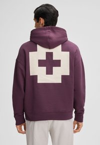 Mann trägt einen lila Hoodie mit einem großen weißen, pixelartigen Kreuz auf der Rückseite, kombiniert mit hellen Hosen.