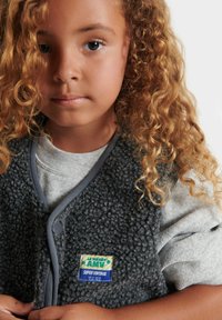 Fleece vesta v tmavě šedé s šedým lemem, s obdélníkovou nášivkou. Nositá přes světle šedou dlouhou rukávovou košili s jemnou texturou.