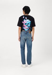 Black t-shirt with colorful skull and butterflies graphic on back, paired with blue denim jeans and white sneakers. Back view shown.