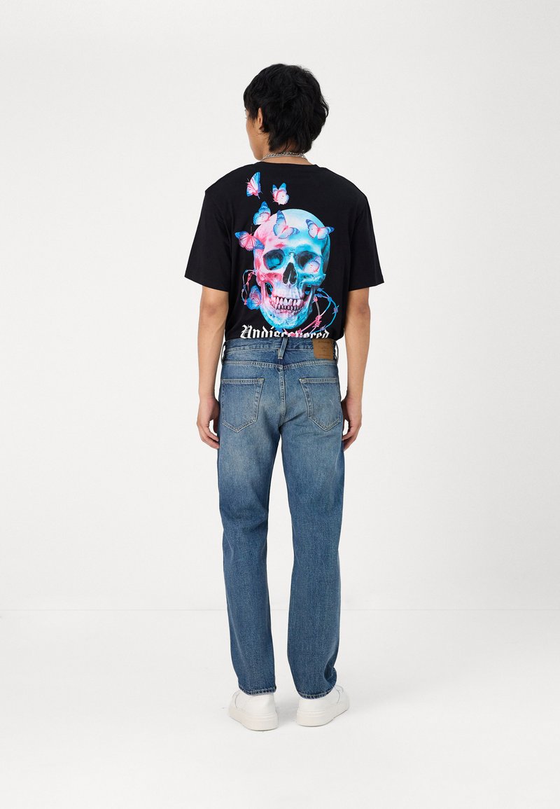 Camiseta negra con un gráfico colorido de calavera y mariposas en la espalda, combinada con jeans de mezclilla azules y zapatillas blancas. Vista trasera mostrada.