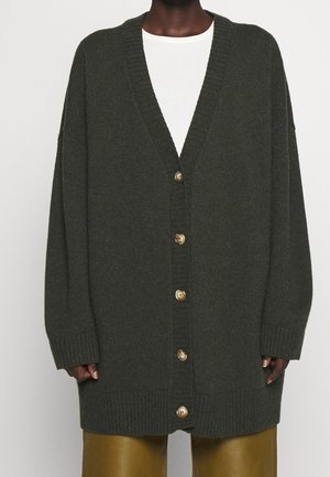 Cardigan oversize boutonné de couleur olive foncé porté par-dessus une chemise blanche avec un pantalon en cuir marron, vu du cou jusqu'à mi-cuisse.