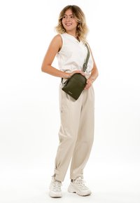 Jauna sieviete stāv, valkājot bezroku baltu kreklu, bēšas bikses, baltus sporta apavus un tur mazu olīvu zaļu krustenisko somu.