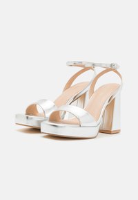 Silver high-heel sandals with a platform, open toe design, and ankle strap. Metallic finish and block heel complement the overall style.