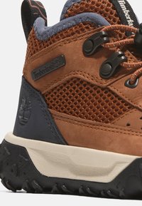 Scarpa da trekking in suede marrone e mesh con suola in gomma nera, design con lacci e etichetta con logo Timberland. Presenta accenti rinforzati e mesh traspirante.