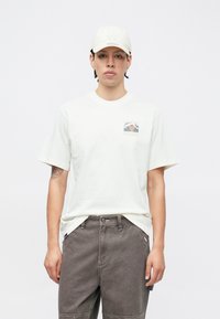 Kreemjas lühikeste varrukatega T-särk, mille vasakul rinnataskul on mägimaastiku kujutis, koos tumehallide cargo-põlvikutega.