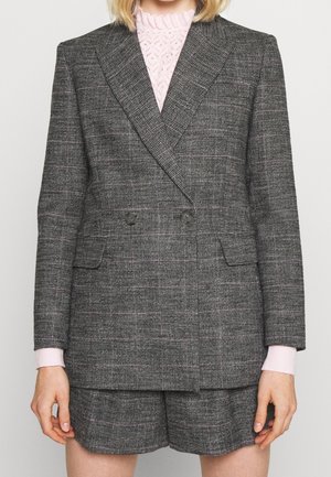 Osoba ubrana w szarą kratowaną dwurzędową marynarkę z dopasowanymi szortami oraz różowy koronkowy top z wysokim kołnierzem pod spodem.