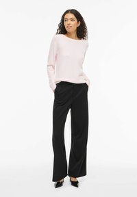 Femme aux cheveux bouclés portant un pull à manches longues rose clair, un pantalon noir à jambes larges et des talons pointus noirs, debout les mains dans les poches.