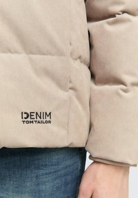 Manga de chaqueta acolchada beige con el logo "DENIM TOM TAILOR" en bordado negro sobre unos vaqueros de mezclilla azul y mano visible.