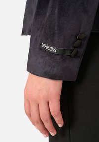 Mörkblå sammetskavajärmsärm med tre svarta knappar och en etikett med "OPPOSUITS"-logotypen. En hand visar lätt hudton och vilar på tyget.