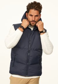 Marineblaue, gesteppte Weste mit Kapuze, Reißverschluss, Seitentaschen und einem darunter getragenen strukturierten Strickpullover. Ein Mann richtet den Kragen der Weste.