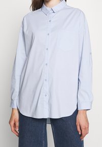 Woman wearing a loose-fitting light blue button-up shirt with long sleeves and a chest pocket, paired with dark blue jeans.