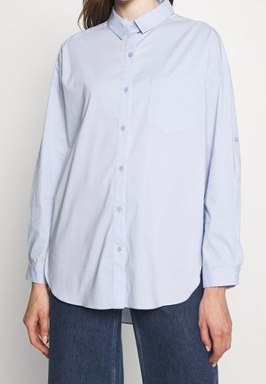 Mujer vistiendo una camisa azul claro de botones, holgada, con mangas largas y un bolsillo en el pecho, combinada con jeans azul oscuro.