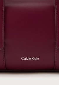 Bordo kožna torba s glatkom teksturom i srebrnim logom „Calvin Klein“. Struktuiran dizajn s naglascima šivanja.