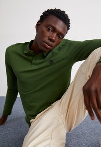 Camisa polo verde de manga longa com colarinho e dois botões, apresentando um pequeno logotipo. Combinada com calças texturizadas de cor clara.