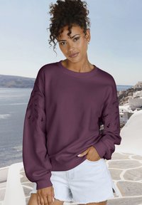 Sudadera burdeos con diseño de recortes intrincados en las mangas, puños y dobladillo acanalados, combinada con pantalones cortos de mezclilla claros.