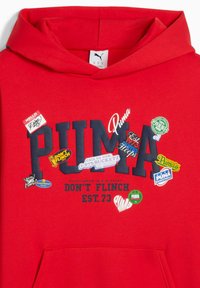 Lys rød hættejakke med "PUMA" i marineblå, dekoreret med forskellige farverige klistermærker og slogans, inklusive "DON'T FLINCH" og "EST. 73."