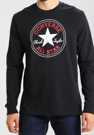 Person trägt ein schwarzes Langarmshirt mit einem roten und weißen Converse All Star Chuck Taylor Logo auf der Brust.