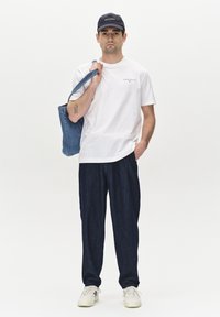 Bela bombažna majica z majhnim logotipom, temno modri jeans hlače, denim torba, črna kapa in bele superge, stoji proti enotni ozadju.