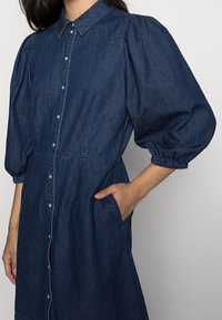 Denim dress in dark blue with a collared neckline, puffed sleeves, button-down front, and side pockets. Features gathered cuffs and seam details.