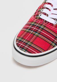 Rode geruite canvas sneaker met witte veters en rubberen zool, met een gestructureerde neuskap en contrasterende zwarte rand langs de rand.