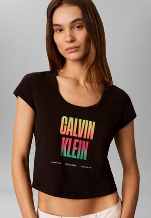 Młoda kobieta z prostymi brązowymi włosami, ubrana w czarny krótki t-shirt z wielokolorowym napisem "Calvin Klein" z przodu, jasne tło.
