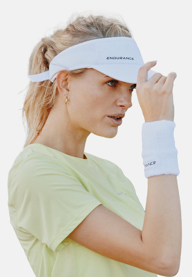 Witte sportvizier met netpanelen, voorzien van het "ENDURANCE"-logo. Vergezeld van een witte polsband. Gekleed met een lichtgroen sportshirt.