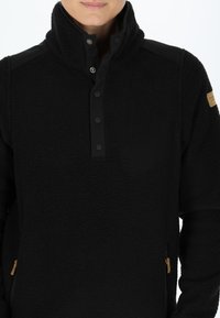 Pullover in pile nero con colletto alto, bottoni a pressione e tasche laterali. Presenta un accento in tessuto liscio e una piccola toppa con logo.