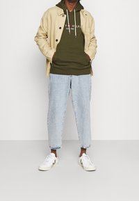 Hombre con una chaqueta beige sobre una sudadera verde oliva con texto, jeans cortos de color azul claro y zapatillas blancas con acentos marrones.