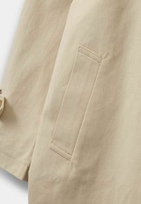 Szczegół beżowej tkaniny bawełnianej z widoczną naszywaną kieszenią. Gładka tekstura i staranne szwy podkreślają prosty design.