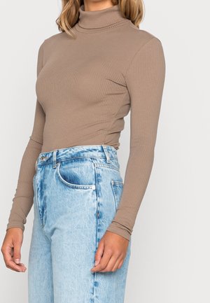 Kobieta ubrana w dopasowany beżowy prążkowany golf i jasnoniebieskie, wysokiego stanu jeansy denim na białym tle.