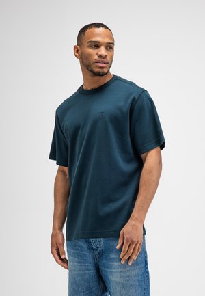 Man wearing dark teal short-sleeve t-shirt and blue jeans, looking to his left against plain light gray background.