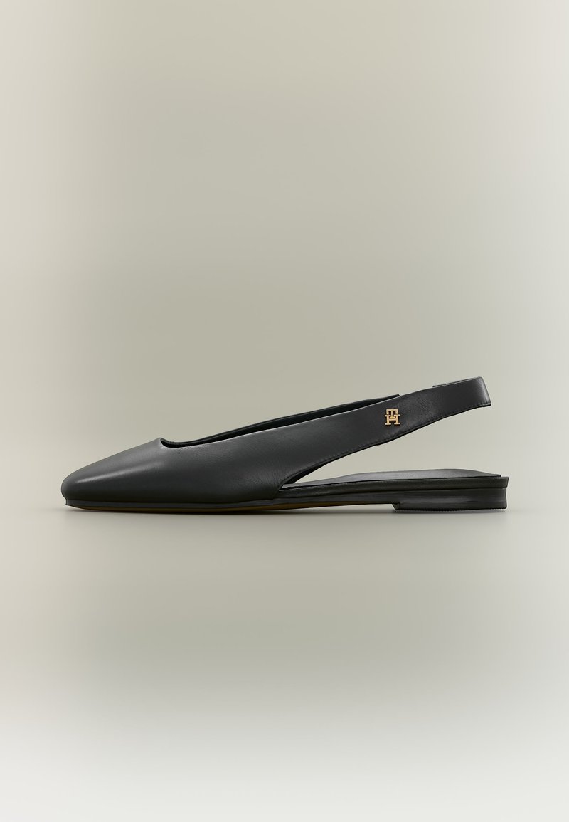 Μαύρο δερμάτινο πάνινο παπούτσι με λουράκι, μυτερή μύτη και χρυσή λογότυπο διακόσμηση στο λουράκι, παρουσιάζεται σε ουδέτερο φόντο.