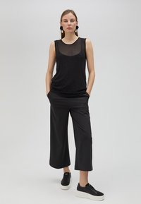 Svart ärmlös topp med en genomskinlig panel och subtila detaljer, parat med vida svarta byxor och svarta slip-on skor.