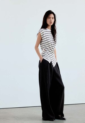 Femme aux longs cheveux foncés portant un haut sans manches blanc et noir rayé et un pantalon large noir, debout contre un mur uni.