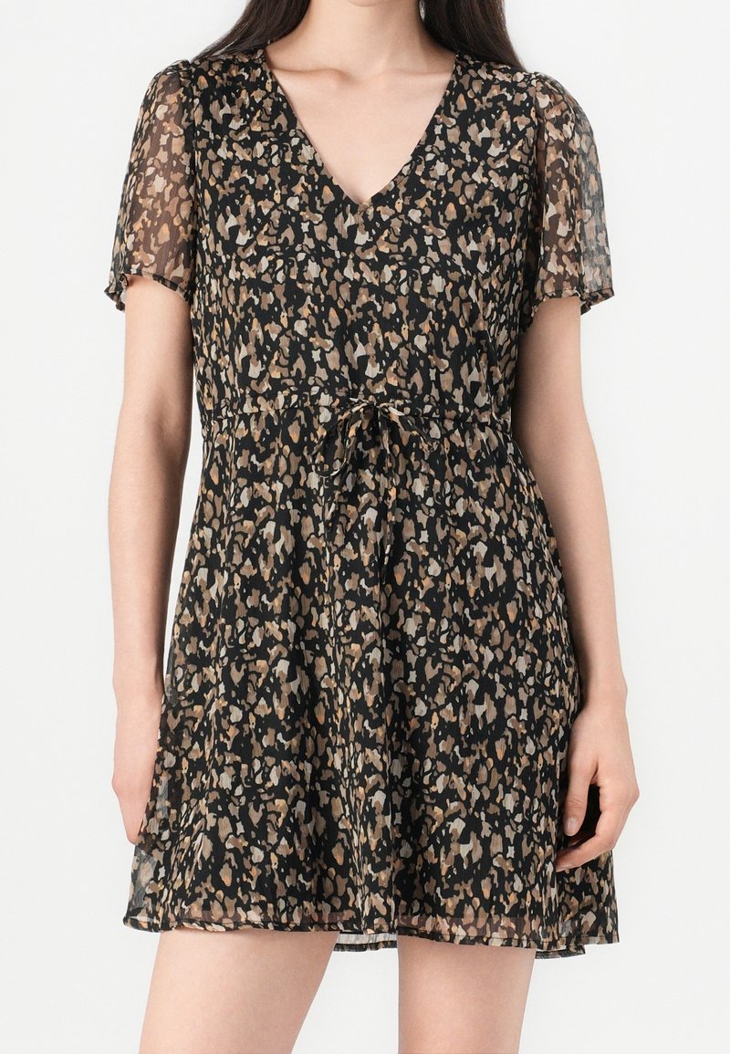Femme portant une robe noire à col en V avec de courtes manches transparentes et des taches abstraites marron et beige, ceinturée à la taille avec un cordon.