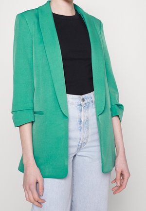 Persoon in een felgroene blazer met opgerolde mouwen over een zwart shirt en lichtblauwe high-waisted jeans tegen een egale achtergrond.