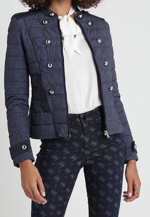 Kobieta w granatowej pikowanej kurtce z metalowymi guzikami na białej bluzce z kokardą, w połączeniu z granatowymi wzorzystymi spodniami.