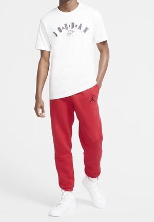 Mann trägt weißes Jordan Air T-Shirt, rote Jordan Sweatpants mit Logo und weiße Turnschuhe, steht mit einer Hand in der Tasche.
