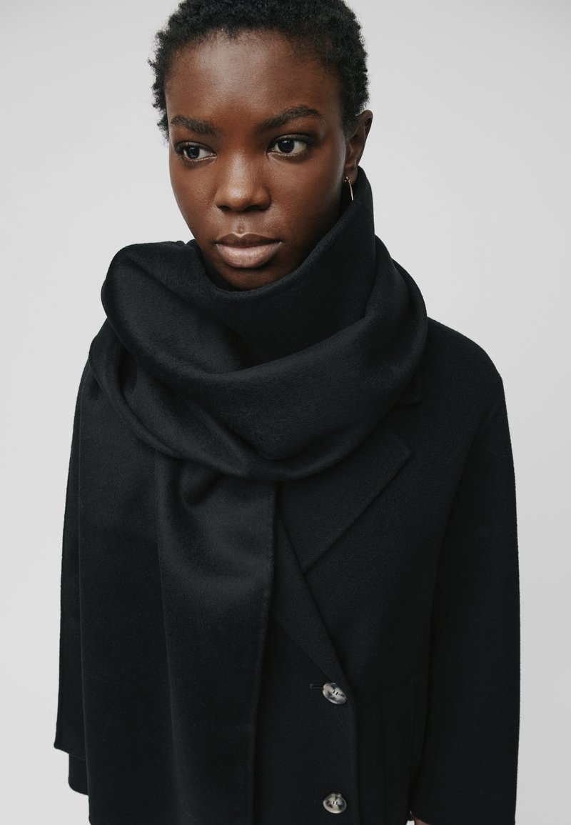 Woman with short curly hair wearing a black coat and large black scarf wrapped around her neck against a plain background.