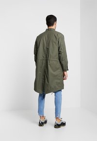 Giacca lunga verde oliva con vita stretta, colletto alto e polsini elastici; realizzata in materiale leggero. Indossata sopra jeans blu e sneaker.